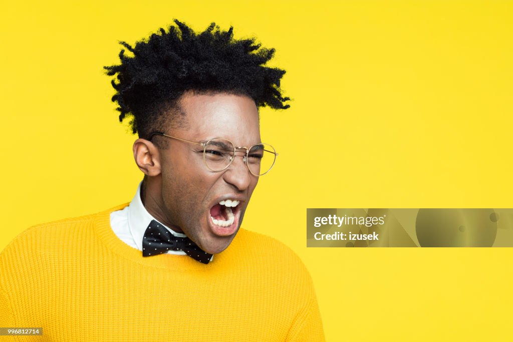 Retrato de hombre joven nerd enojado gritando contra el fondo amarillo