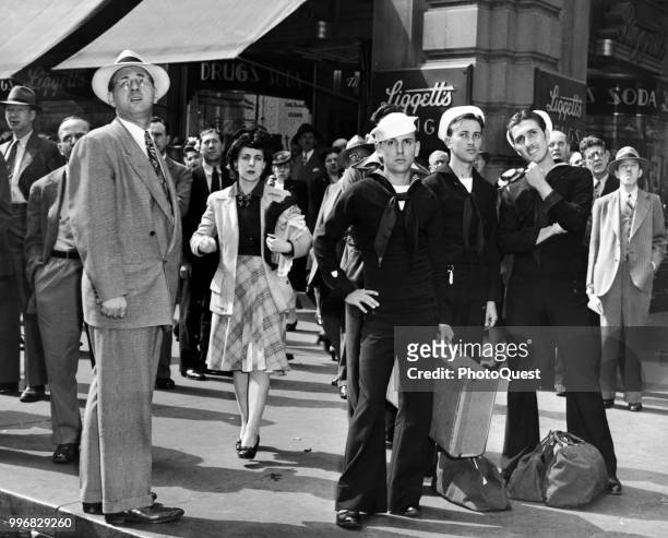 View of a trio of sailors and a large group of civilians as they stand in front of Liggett's Drugs on a Times Square street corner, New York, New...