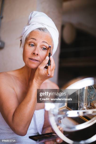 beautiful senior woman putting on make-up and applying eyeshadow - only mature women stock pictures, royalty-free photos & images