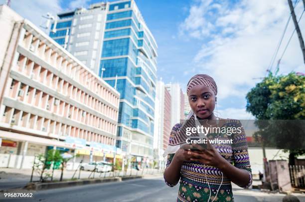 jeune fille africaine écouter de la musique sur les écouteurs de téléphone mobile dans le centre de la ville, regardant la caméra - dar photos et images de collection