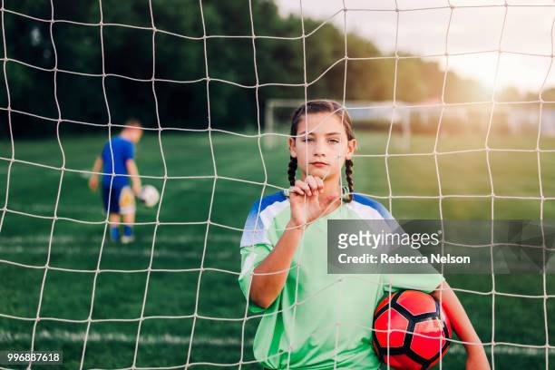 11 year old girl holding soccer ball looking through soccer goal net - mannschaftsfußball stock-fotos und bilder