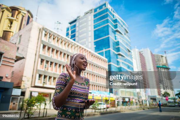 jeune fille africaine écouter de la musique sur les écouteurs de son téléphone intelligent en l’agitant de centre ville - dar photos et images de collection