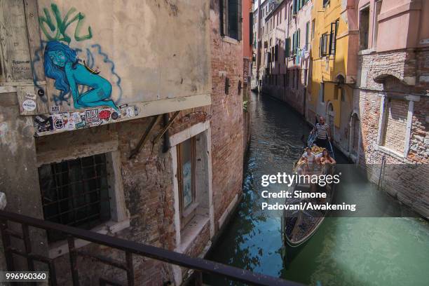 Gondola sails under the bridge of Salizada San Giovanni Grisostomo where there is a graffiti on the wall representing a blue naked woman with a...