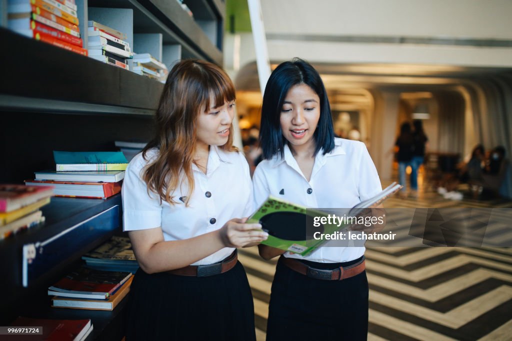 Zwei junge asiatische Studenten in Uniformen im Gespräch in der Campusbibliothek nach Klassen