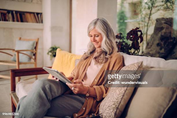 lachende senior vrouw met grijze haren met behulp van tablet pc thuis - blogger woman stockfoto's en -beelden