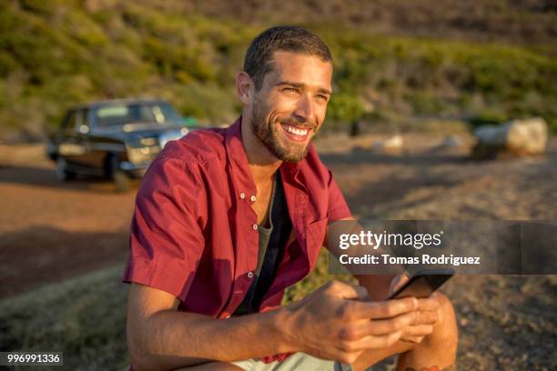 smiling young man with cell phone sitting at the coast with car in background - mobile phone and adventure stockfoto's en -beelden