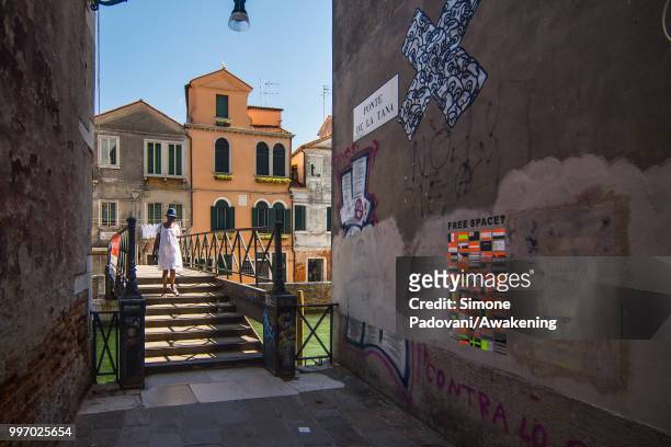 Tourist crosses De La Tana bridge close to Arsenale entrance of Biennale of Venice, with the wall covered by graffiti and tags, on the way to...