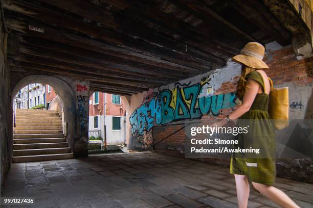 Tourist walks to cross the Anatomia bridge, where there are graffiti and tags on the walls, through Santa Croce district, on July 12, 2018 in Venice,...