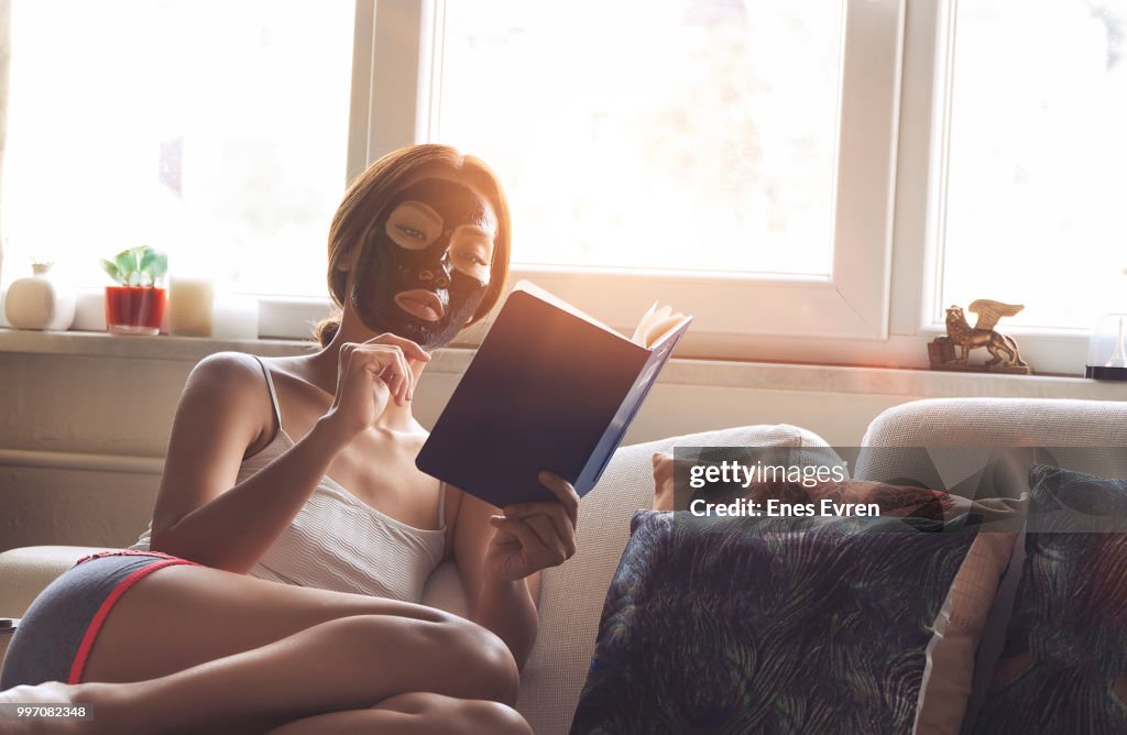 Woman with charcoal face mask reading book (erased designs on pillows)