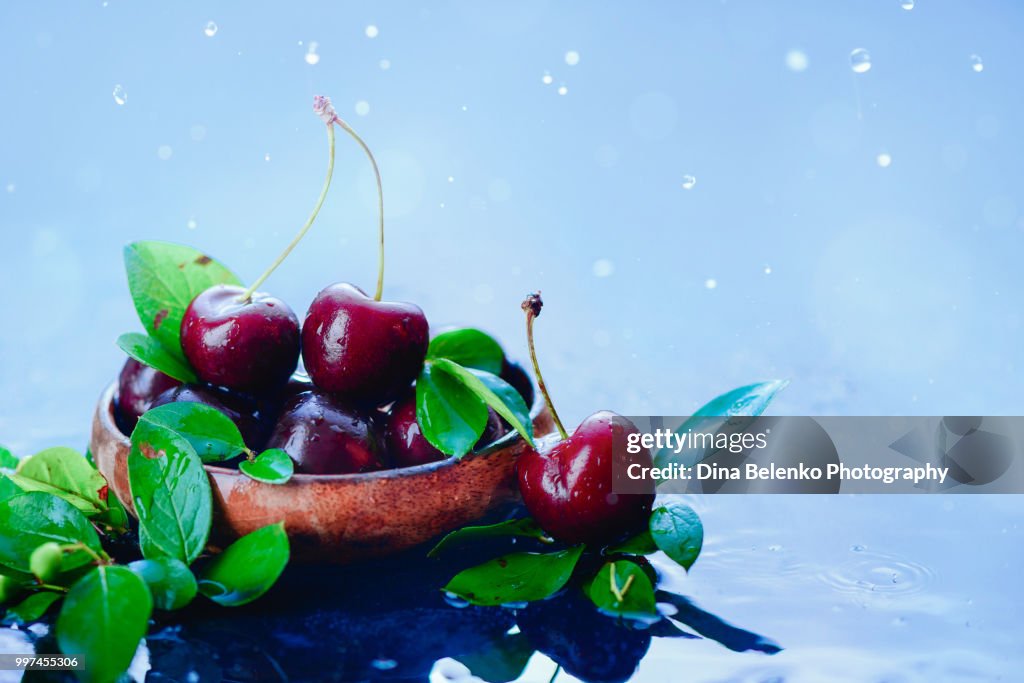 Ripe cherries in a wooden bowl with green leaves on a light wet background with water drops. Harvest concept with copy space. Autumn rain still life.