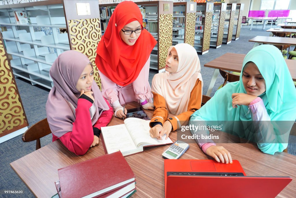 Beautiful young Muslimah students from Southeast Asian with hijab looking to the laptop and having discussion in the library.