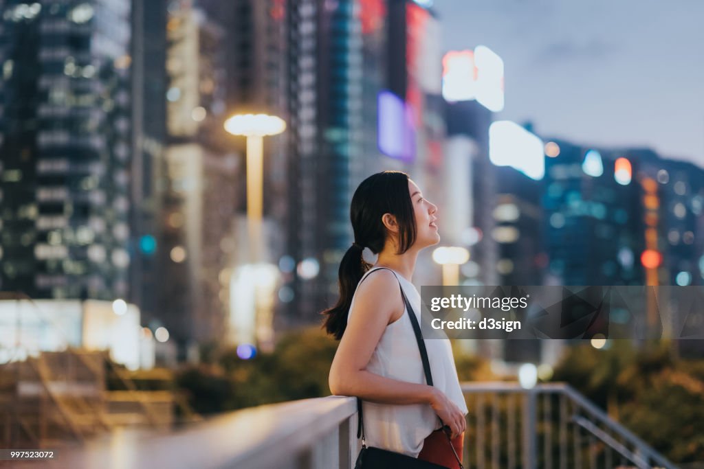 Beautiful young woman resting by the fence and looking up to the sky enjoying the spectacular sunset in city