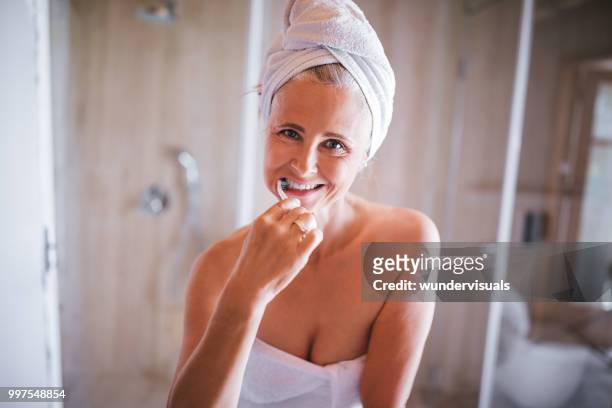 senior woman in bath towels brushing her teeth in bathroom - woman gray hair mirror stock pictures, royalty-free photos & images