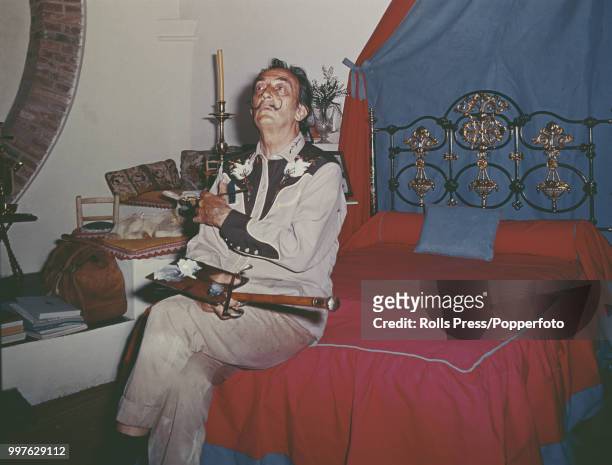 Catalonia born Spanish artist Salvador Dali pictured holding a paintbrush and palette as he sits on a bed in the bedroom of his house near Cadaques...