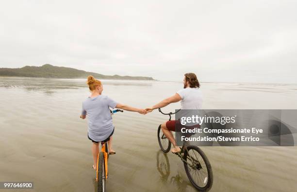 couple holding hands bike riding on beach - dark haired man gray shirt with wine stock pictures, royalty-free photos & images