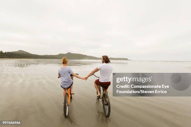couple holding hands bike riding on beach - dark haired man gray shirt with wine stock pictures, royalty-free photos & images