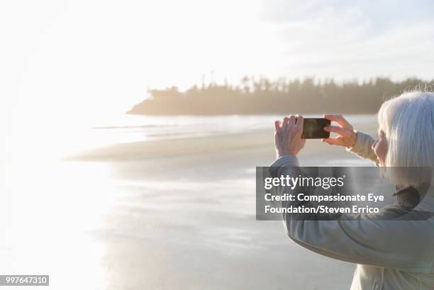 senior woman on beach taking photo of ocean view at sunset - technophiler mensch stock-fotos und bilder