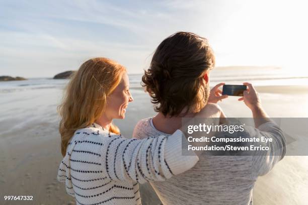 couple on beach taking photo of ocean view at sunset - technophiler mensch stock-fotos und bilder