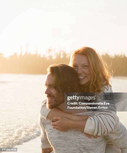 smiling couple piggybacking on beach at sunset - mittelalter mann stock-fotos und bilder