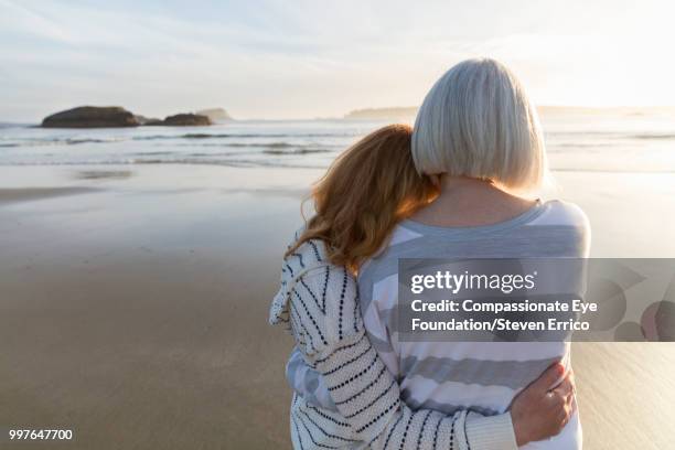 senior woman and daughter hugging on beach looking at ocean view at sunset - femmes de dos enlacée photos et images de collection