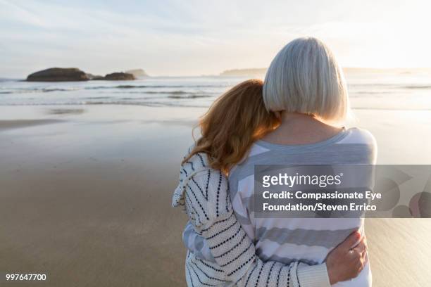 senior woman and daughter hugging on beach looking at ocean view at sunset - head on shoulder stock pictures, royalty-free photos & images