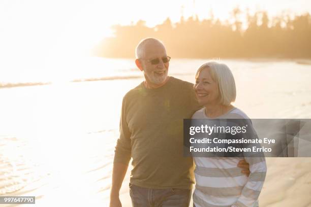 smiling senior couple walking on beach at sunset - canadian pacific women stock pictures, royalty-free photos & images