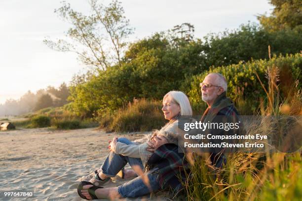 senior couple sitting on beach at sunset - eskapismus stock-fotos und bilder