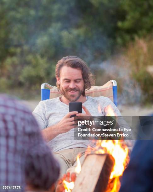 smiling man using cell phone on beach by camp fire - technophiler mensch stock-fotos und bilder