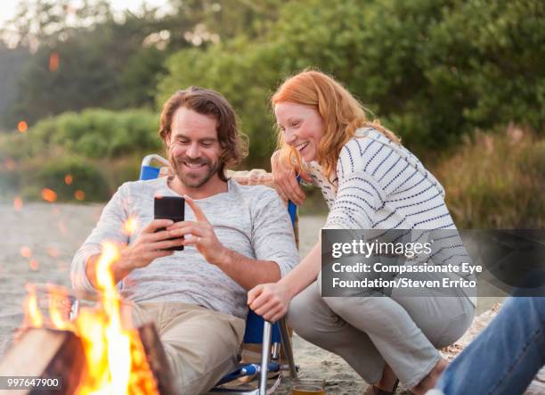 smiling couple using cell phone on beach by camp fire - technophiler mensch stock-fotos und bilder
