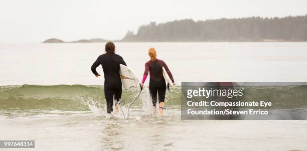 surfers running into waves with surfboards - "compassionate eye" fotografías e imágenes de stock