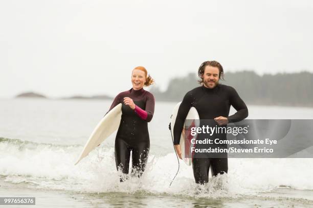 surfers carrying boards in waves - "compassionate eye" fotografías e imágenes de stock