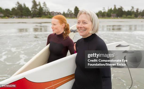 female surfers carrying boards walking along beach - "compassionate eye" fotografías e imágenes de stock