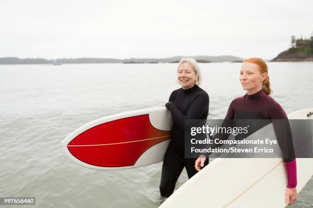 female surfers carrying boards walking along beach - "compassionate eye" fotografías e imágenes de stock