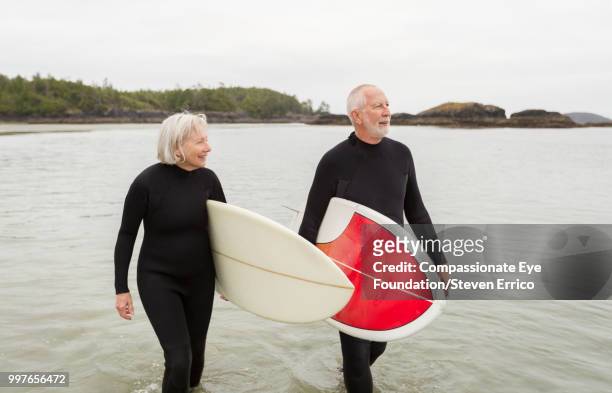 older surfers carrying boards walking along beach - "compassionate eye" fotografías e imágenes de stock