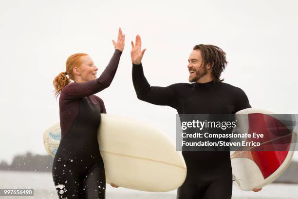 surfers carrying boards high fiving - compassionate eye foto e immagini stock