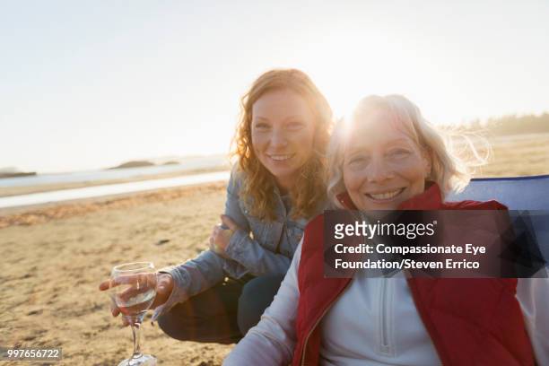 senior woman and adult daughter relaxing on beach at sunset - compassionate eye foundation stock-fotos und bilder