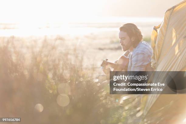 man camping playing guitar on beach at sunset - compassionate eye foundation stock-fotos und bilder