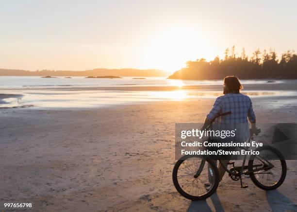 man siting on bike looking at ocean view at sunset - compassionate eye foundation stockfoto's en -beelden