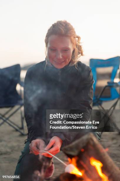 woman toasting marshmallow on camp fire at beach - compassionate eye foundation ストックフォトと画像