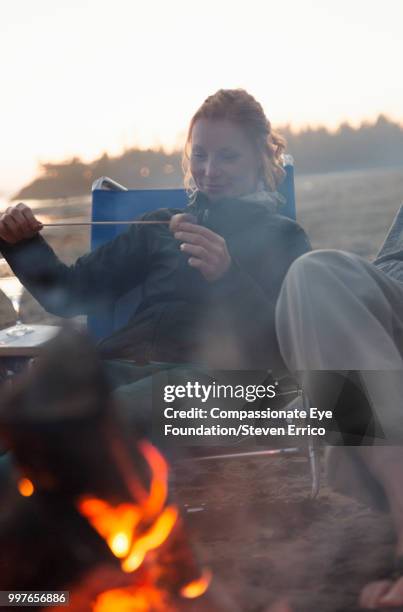 woman eating toasted marshmallow by camp fire at beach - compassionate eye foundation stock-fotos und bilder