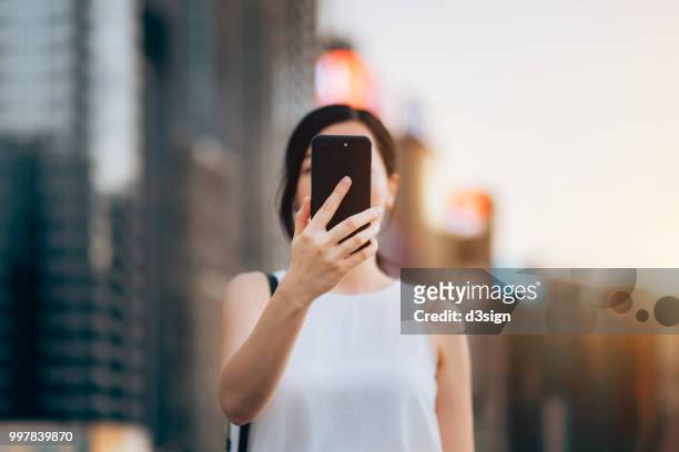 young woman using smartphone outdoors in front of blurry city scene - mensaje de móvil fotografías e imágenes de stock