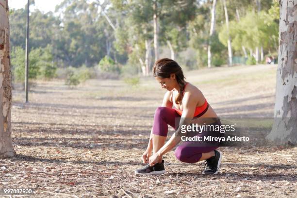 fit, ethnic woman wearing athletic clothing, crouches down to tie her shoes, in a park - mireya acierto stockfoto's en -beelden