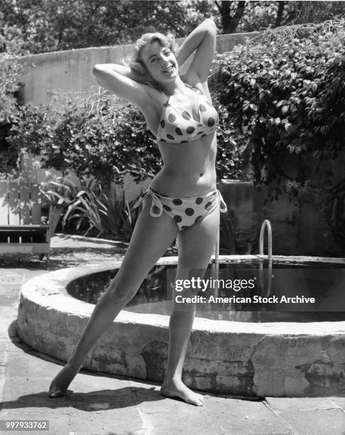 Portrait of an unidentified model, in a two-piece, polka-dot bathing suit, as she poses, arms behind her head, beside a small pool in a backyard, Los...