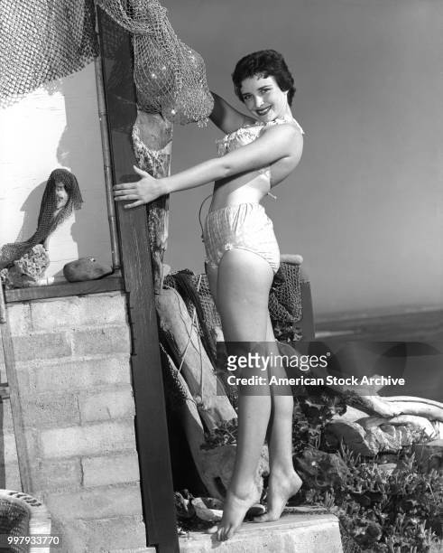 Portrait of an unidentified model, in a two-piece bathing suit, as she poses as the corner of a building draped in fish nets, Los Angeles,...