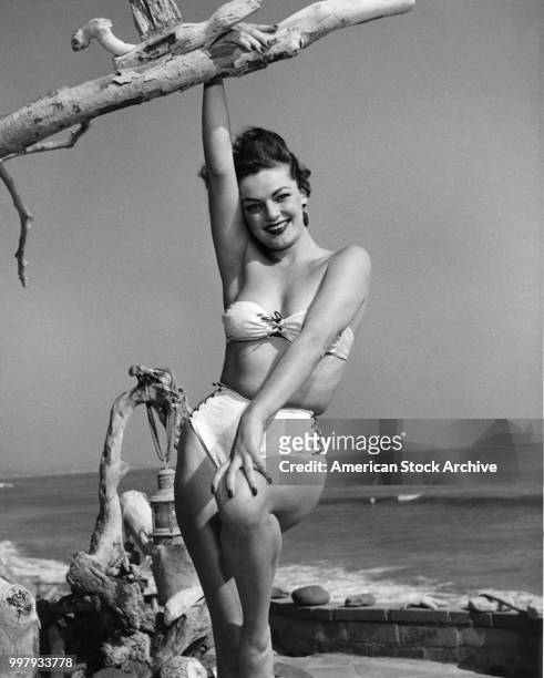 Portrait of an unidentified model, in a strapless, two-piece bathing suit, as she poses, one hand on a piece of driftwood, at the seaside, Los...