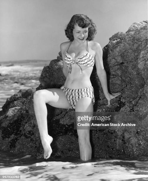 Portrait of an unidentified model, in a two-piece bathing suit, as she stands against rocks, one leg in the ocean surf, Los Angeles, California,...