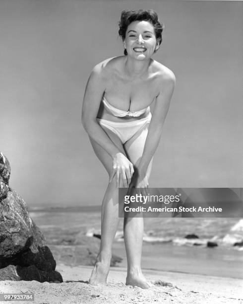 Low-angle portrait of an unidentified model, in a strapless, two-piece bathing suit, as she poses, hands on her knees, at the beach, Los Angeles,...