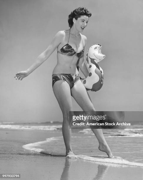 Low-angle portrait of an unidentified model, in a two-piece bathing suit, as she holds an inflatable seahorse under one arm and dips toes in the surf...