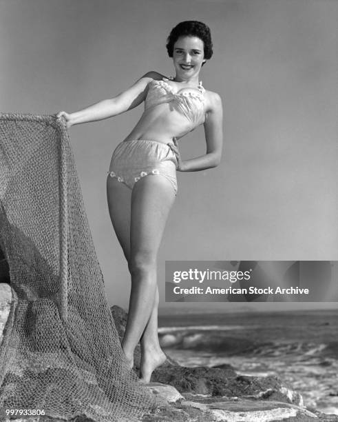 Low-angle portrait of an unidentified model, in a two-piece bathing suit, as she poses, one hand on a fishing net, at the beach, Los Angeles,...