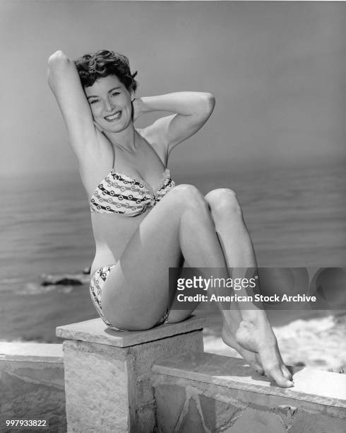 Portrait of an unidentified model, in a two-piece bathing suit, as she sits on a low wall that overlooks the ocean, Los Angeles, California, January...